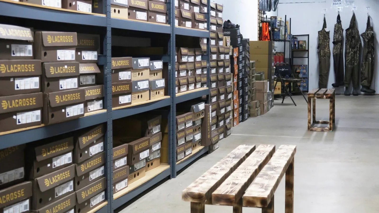 Warehouse interior with tall shelving units labeled "LACROSSE" containing brown and tan boxes, wooden benches in foreground, and tall decorative conifer sculptures on right side.