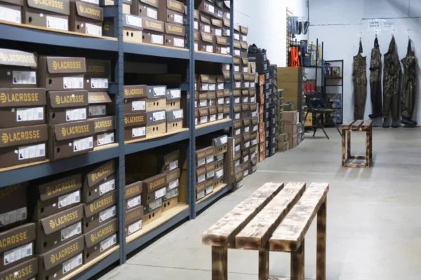 Warehouse interior with tall shelving units labeled "LACROSSE" containing brown and tan boxes, wooden benches in foreground, and tall decorative conifer sculptures on right side.